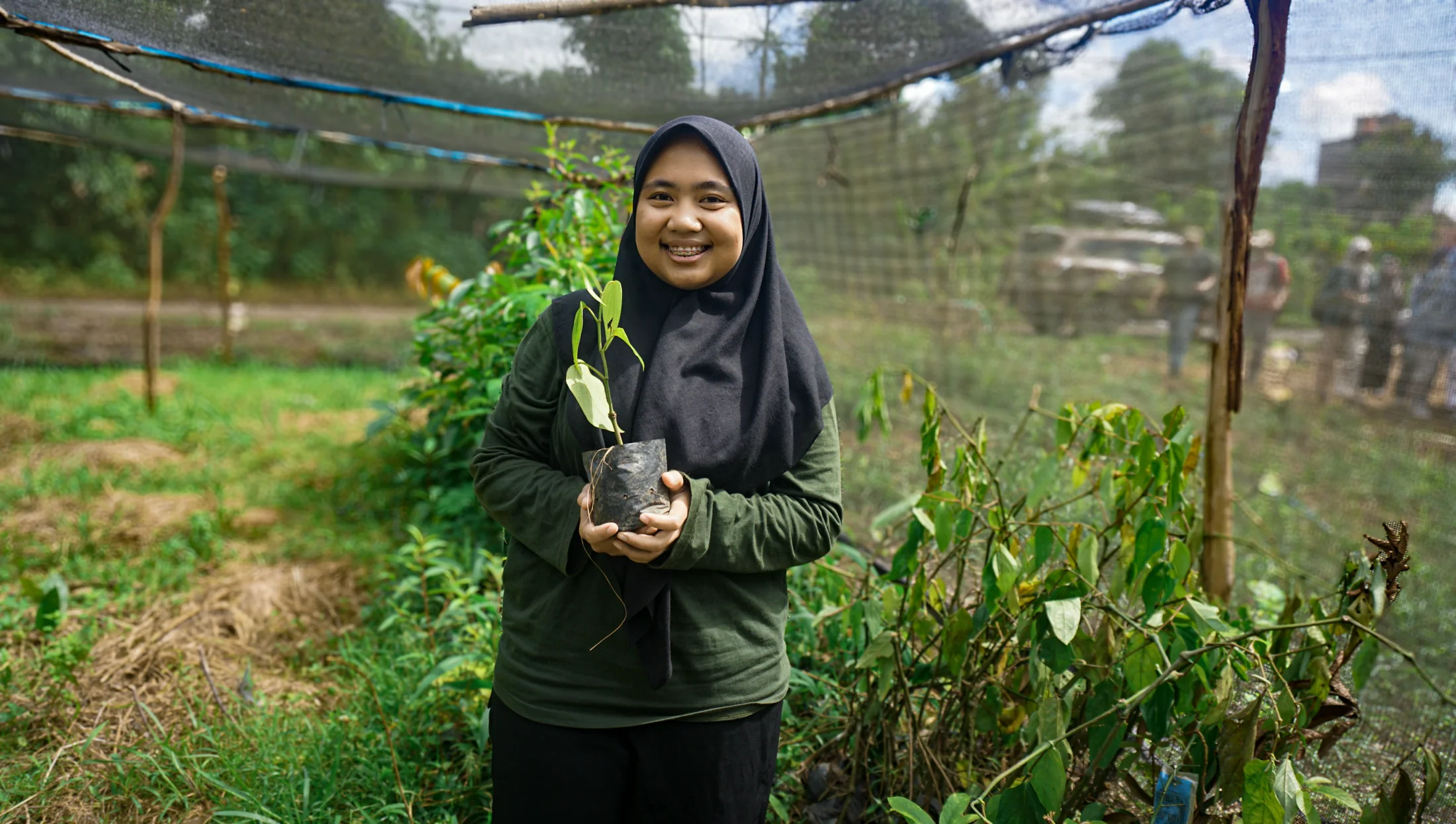 A nursery worker smiling as she holds a healthy young pepper seedling under the shade netting